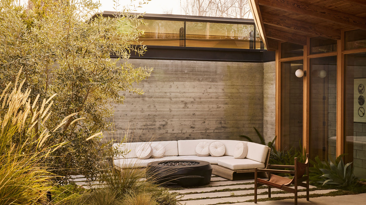 A modern outdoor patio area featuring a curved white sectional sofa, a round black fire pit, a wooden chair, and a concrete wall backdrop with architectural wood and glass details.