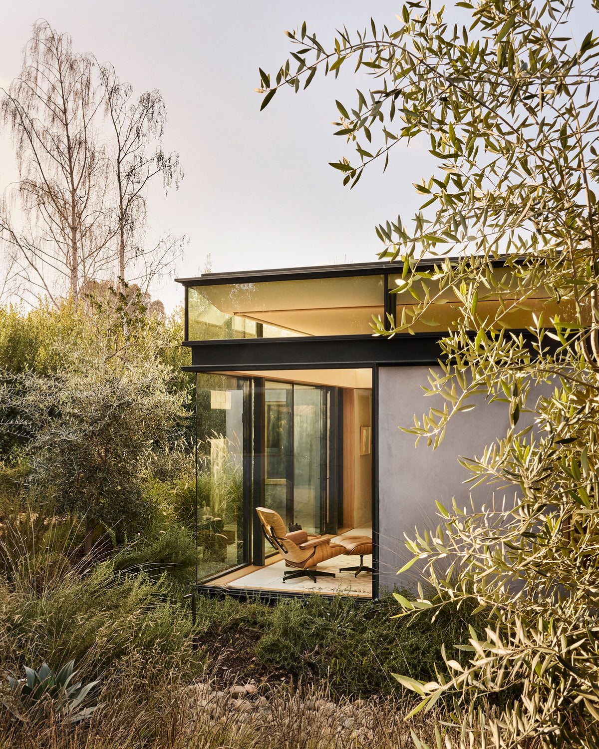 Modern glass house nestled in dense green foliage, featuring a large sliding glass wall and a visible lounge chair inside. The structure has a dark frame and a light gray side wall, set against a soft sky background.