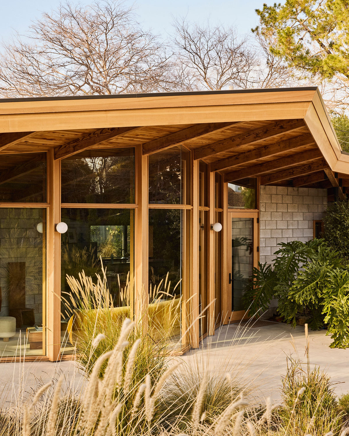 Modern wooden house exterior with large glass windows, warm wood framing, a covered porch, stone wall, and landscaped grasses in the foreground.