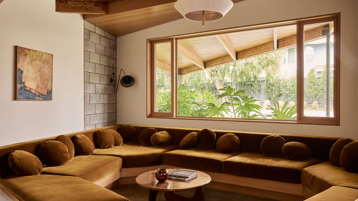 A modern living room featuring a U-shaped sectional sunken sofa upholstered in a warm brown velvet fabric, set in front of a large window with a view of lush green plants outside.