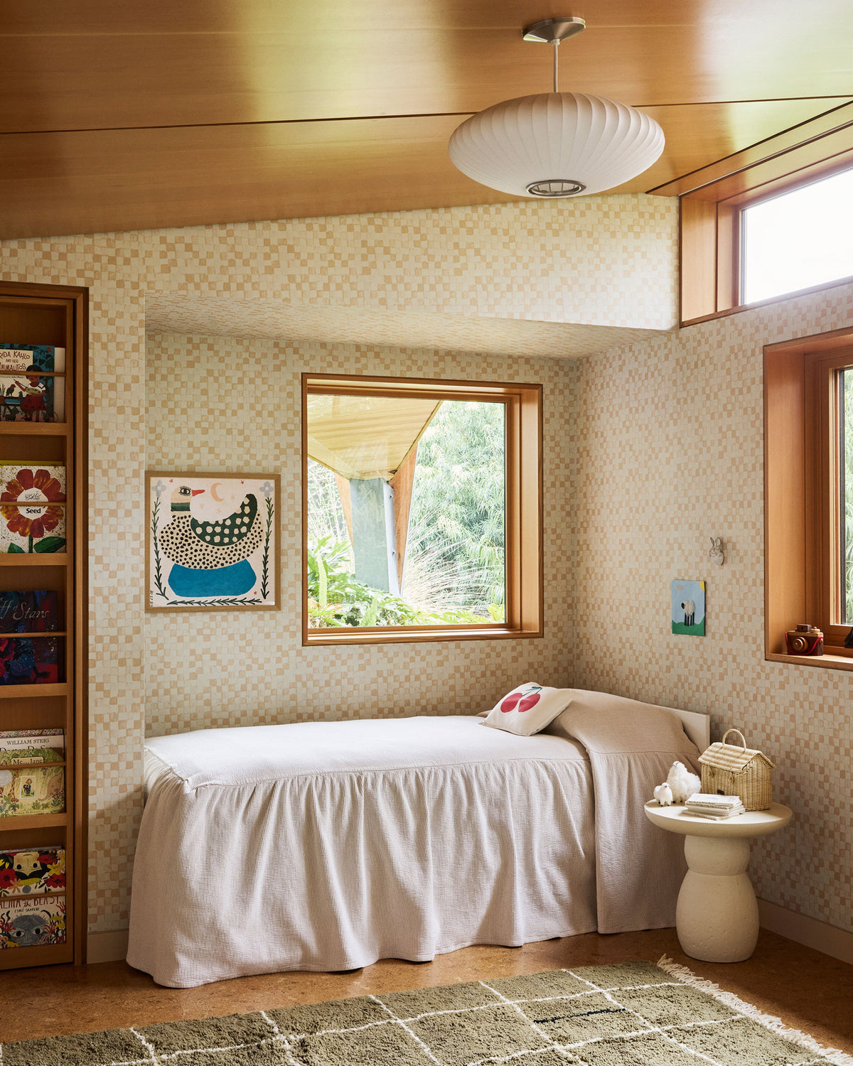A cozy, vintage-style bedroom or nursery featuring a single bed with a white slipcover, patterned wallpaper, a wooden ceiling, a white ribbed pendant light, and a small side table.