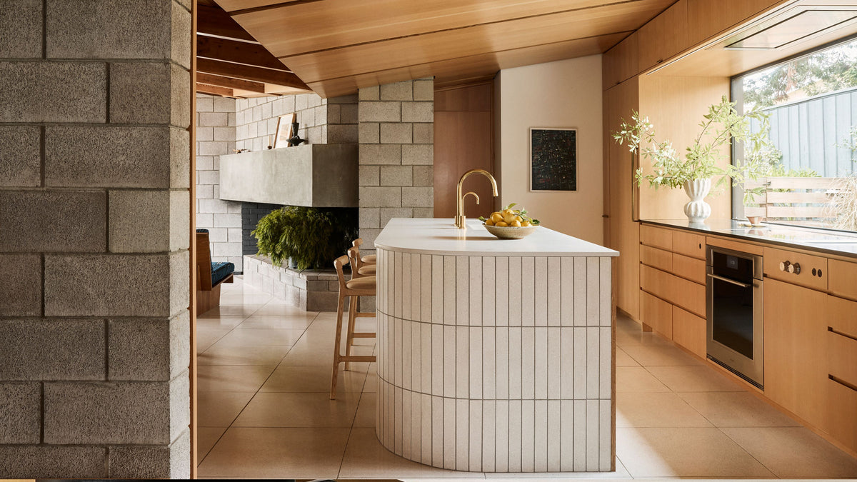 A modern kitchen interior with a central white island featuring vertical wood slats, a gold faucet, a bowl of fruit, light wood cabinetry, a stone wall, and a large window.