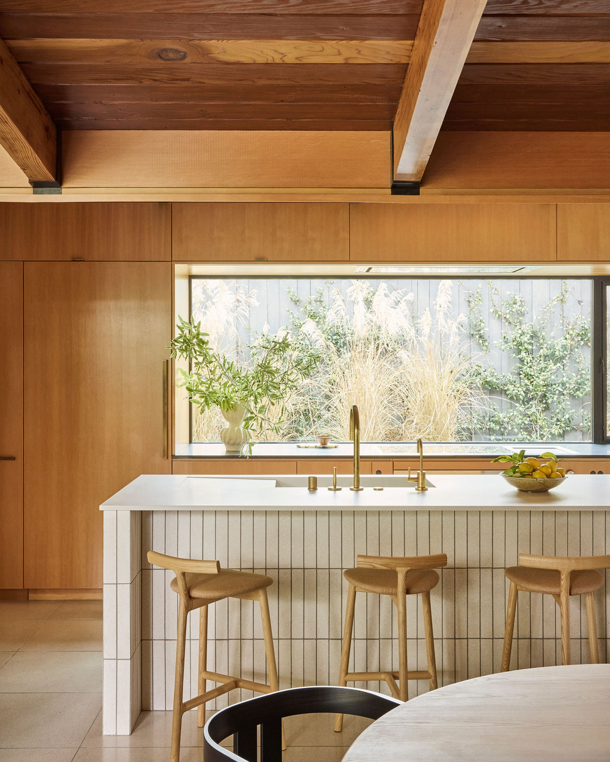A modern kitchen interior with a wood-paneled ceiling, a white tiled island, three wooden bar stools, and a large window looking out onto greenery.