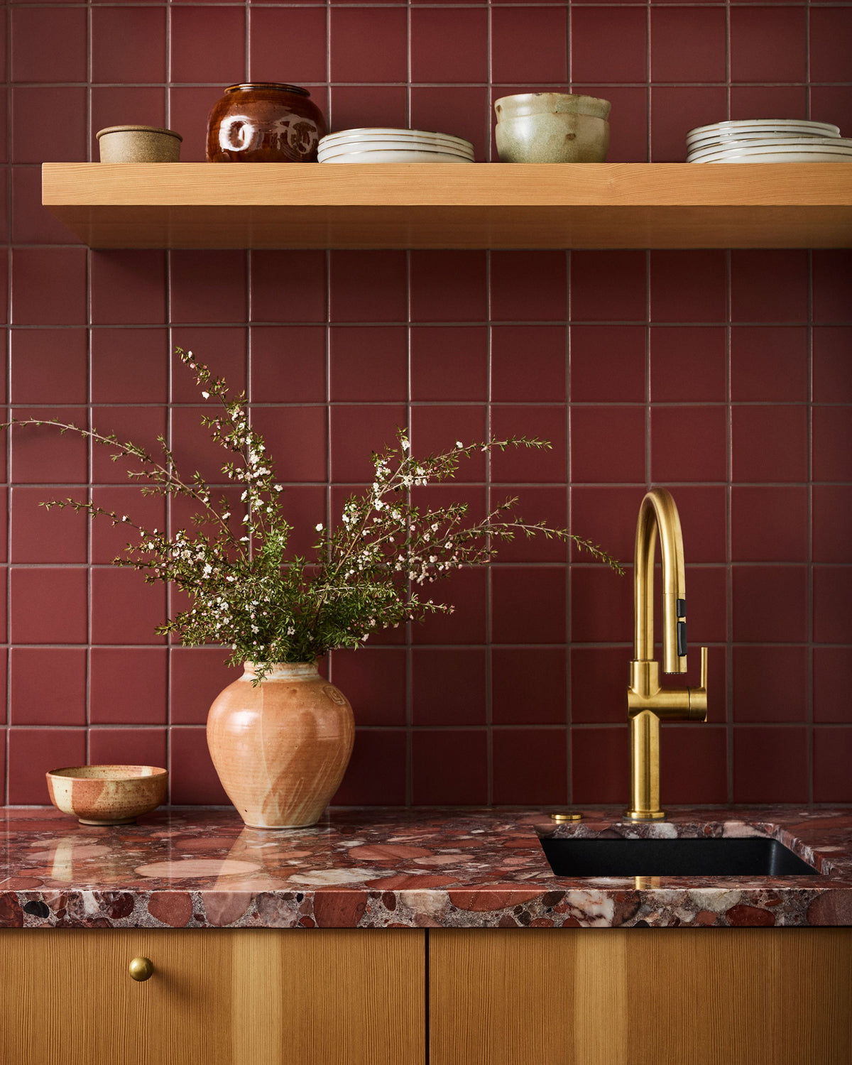 A kitchen scene with a deep red tiled backsplash, a warm wood floating shelf, a red-and-white marble countertop, a black sink, and a tall brushed-gold faucet. A terracotta vase with delicate greenery sits on the counter beside the sink.