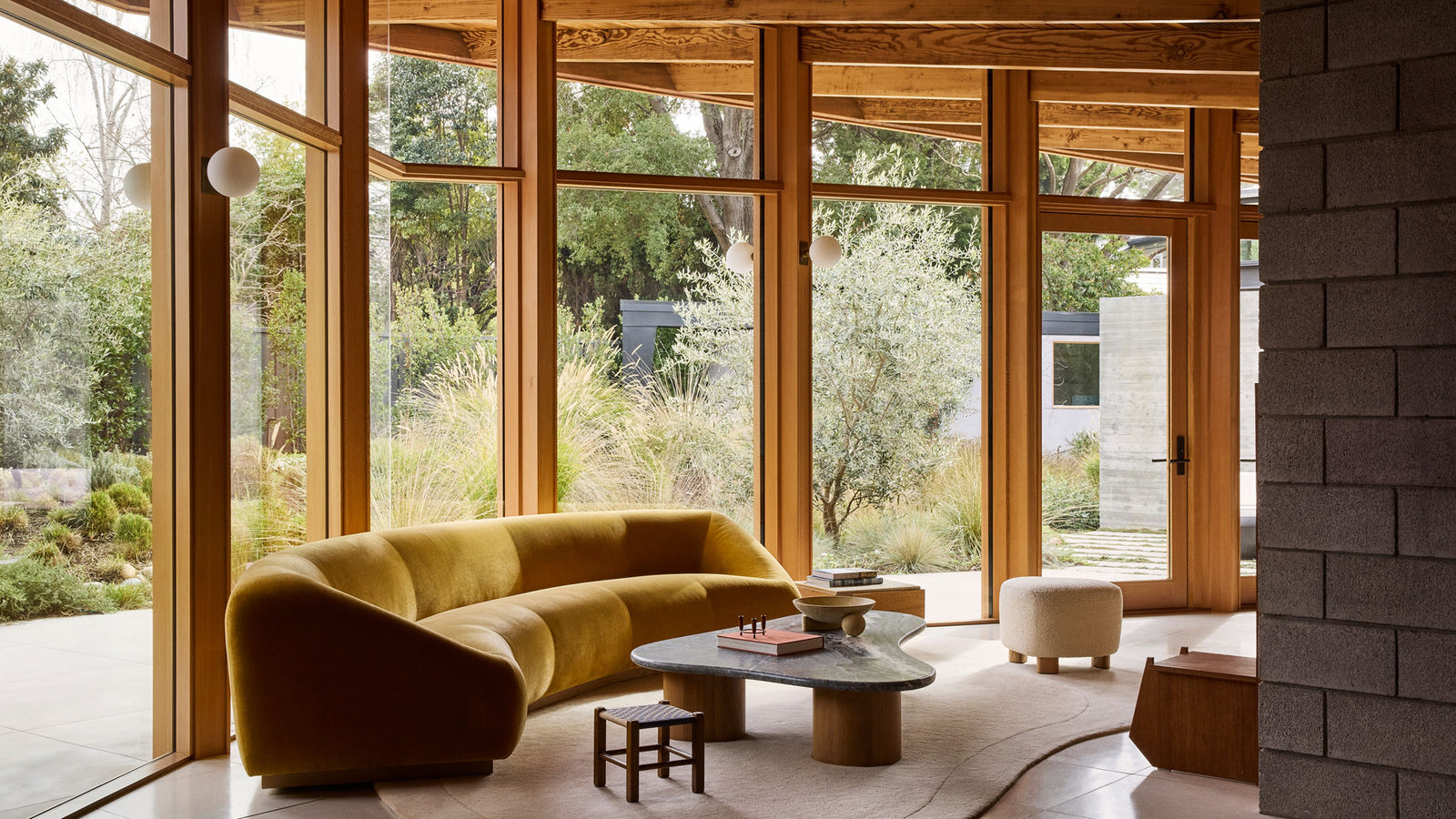 A living room interior with a curved mustard-yellow velvet sofa, a round stone-top coffee table, and large wood-framed windows overlooking a landscaped garden.