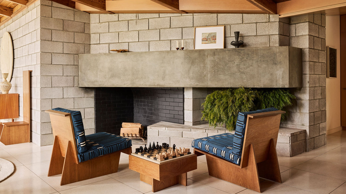 A mid-century modern living room featuring two wooden lounge chairs with blue striped cushions, a matching wooden coffee table with a chess set, and a large concrete fireplace built into a gray brick wall.