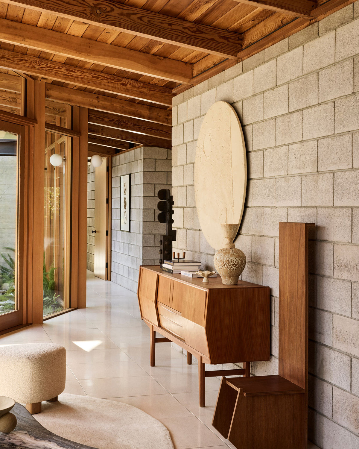 A mid-century modern wooden credenza in a warm medium brown finish, styled in a contemporary home interior with a light gray brick wall, a large oval mirror, and exposed wood ceiling beams.