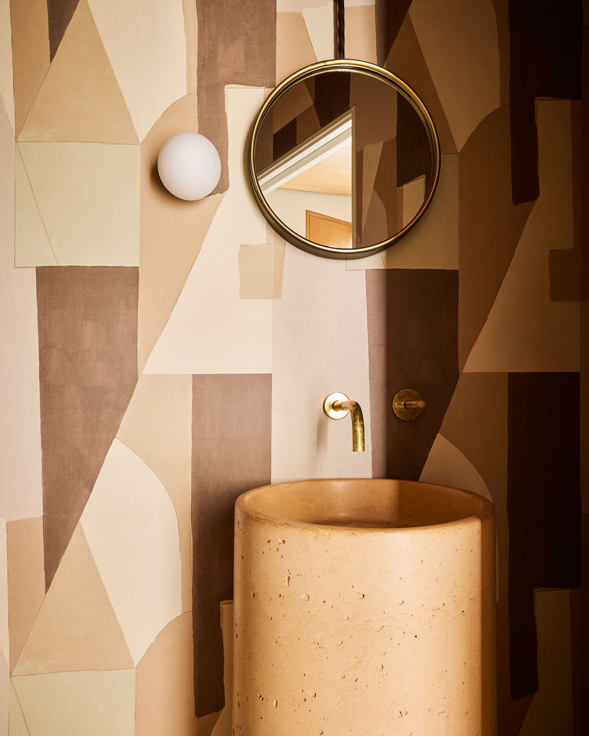 A modern bathroom vanity setup featuring a cylindrical stone pedestal sink, a round gold-framed mirror, and a geometric patterned wall in warm beige, brown, and cream tones.
