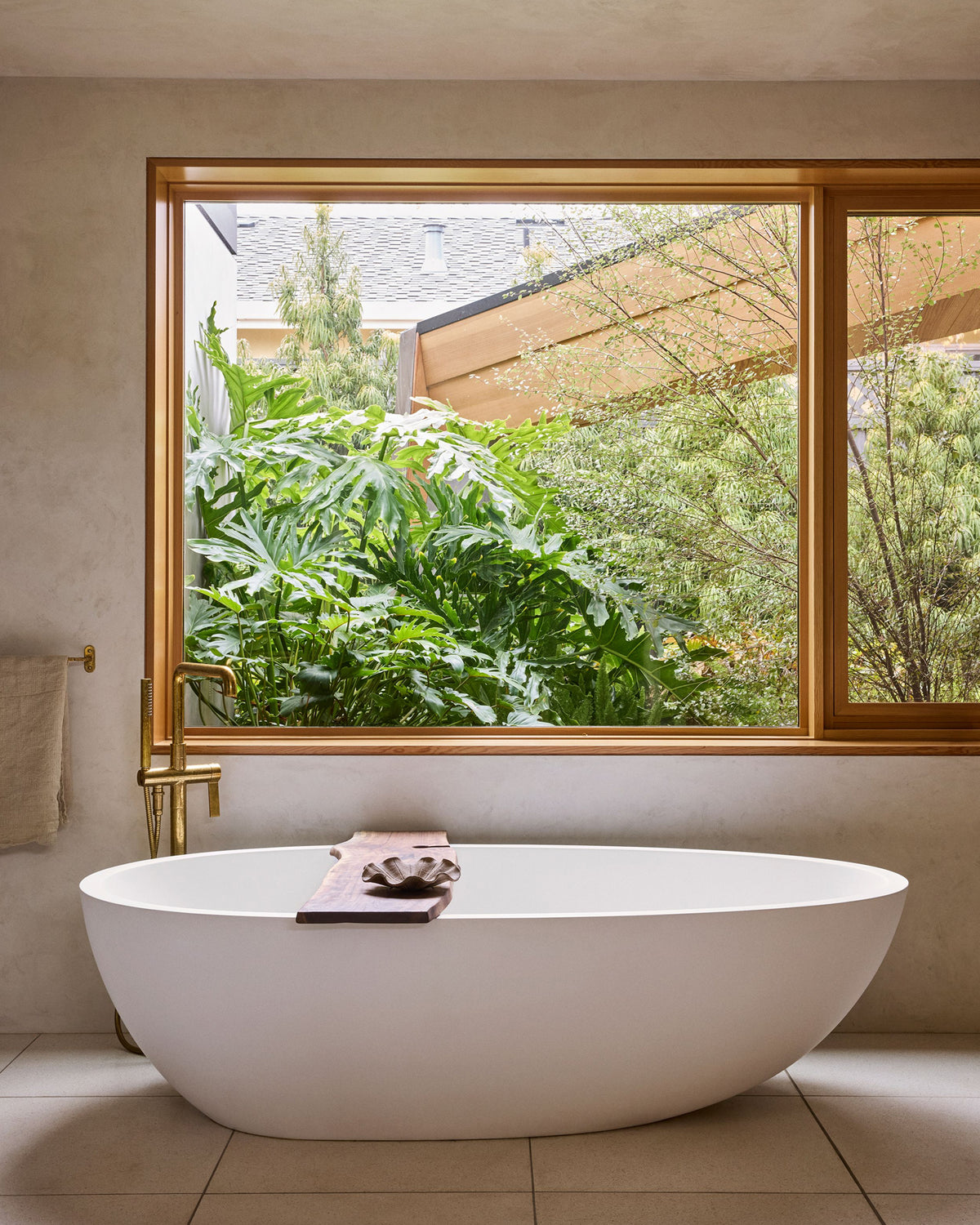 A white freestanding oval bathtub in a modern bathroom with a large window framed in warm wood, overlooking lush green plants and outdoor greenery.