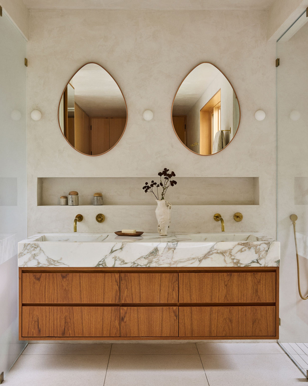 A modern floating bathroom vanity with a white marble countertop, warm wood drawers, and two teardrop-shaped mirrors on a textured beige wall.