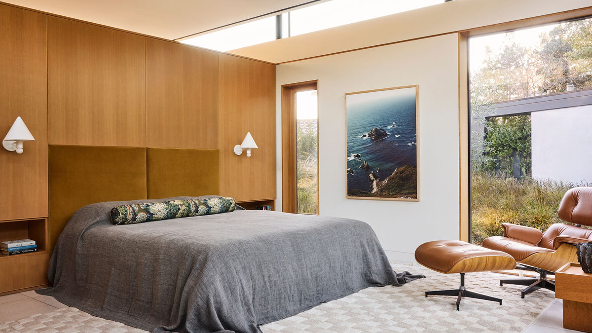 A modern bedroom interior with a bed dressed in gray bedding, a wood-paneled headboard wall, a framed coastal landscape print on the wall, and a brown lounge chair with an ottoman beside large windows.