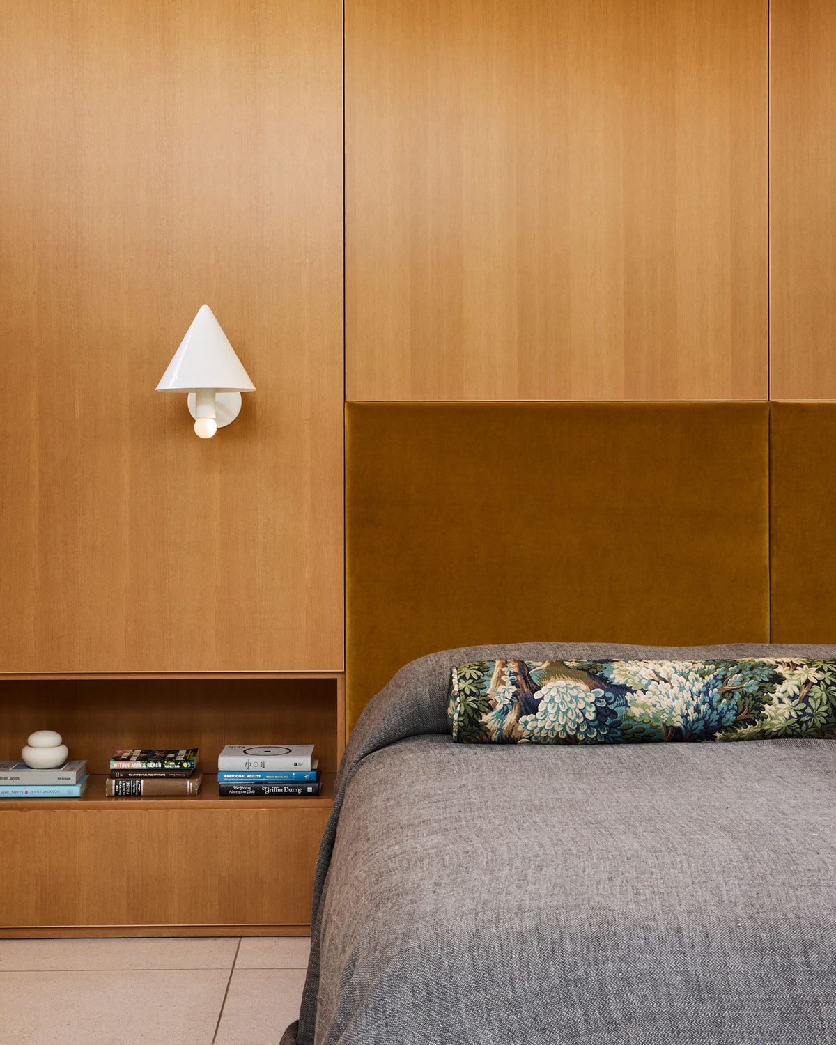 A modern bedroom interior featuring a built-in wooden headboard and shelving, a gray textured bedspread, a decorative floral cylindrical pillow, and a white wall sconce.