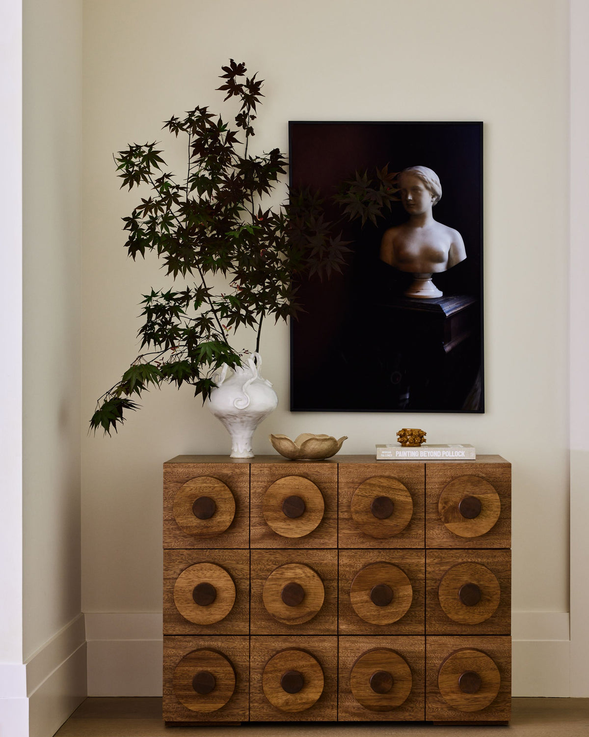 A modern wooden cabinet with 12 round-drawer compartments, styled in a home interior next to a white vase with a leafy plant and a large framed art print on the wall.