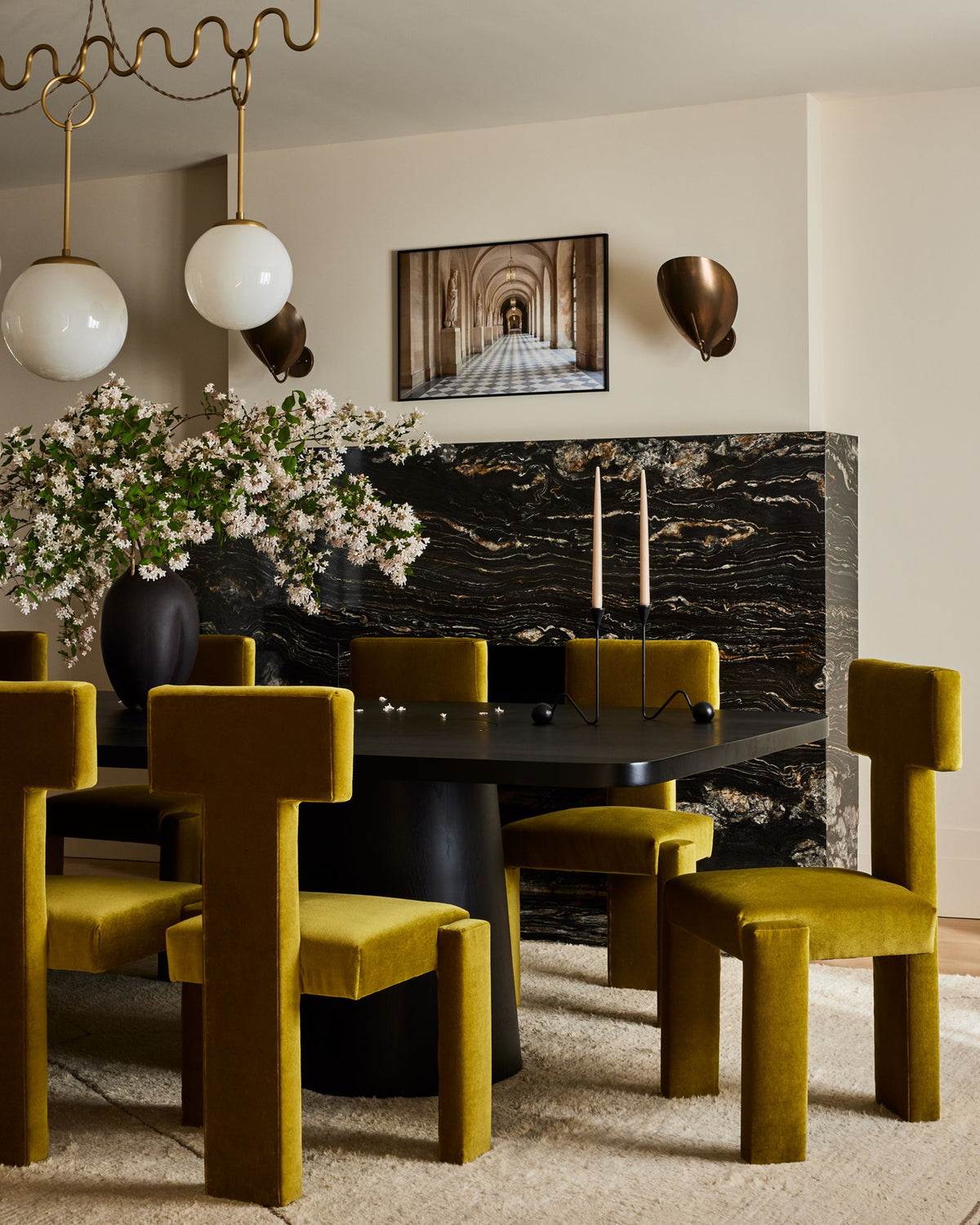 A dining room scene with mustard yellow velvet chairs around a dark round table, set in front of a black marble wall panel and cream walls. Pendant lights, wall sconces, a framed picture, and a floral arrangement are visible.