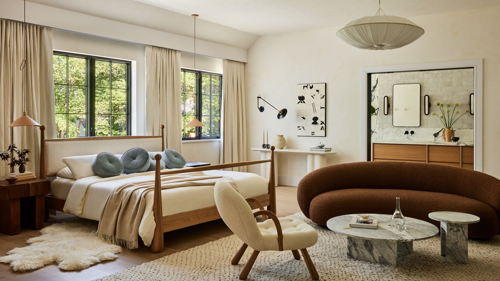 A beautifully styled bedroom interior featuring a wooden bed with cream bedding and blue-gray pillows, a curved brown sofa, a cream accent chair, marble coffee tables, and large windows with beige curtains.