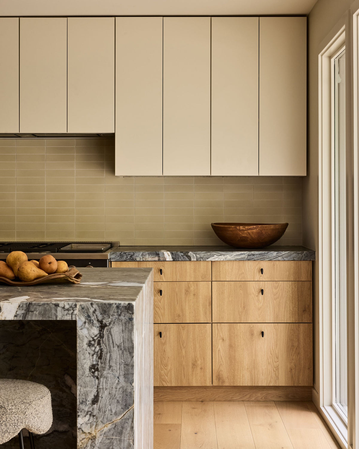 A kitchen interior with cream upper cabinets, light wood lower cabinets, a gray marble countertop and island, a beige tile backsplash, and a copper bowl on the counter.
