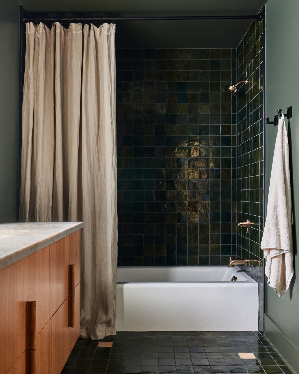 A modern bathroom interior featuring a white bathtub set against a dark green glossy tiled wall, with a beige curtain, a wooden vanity, and a white towel hanging on a black rack.