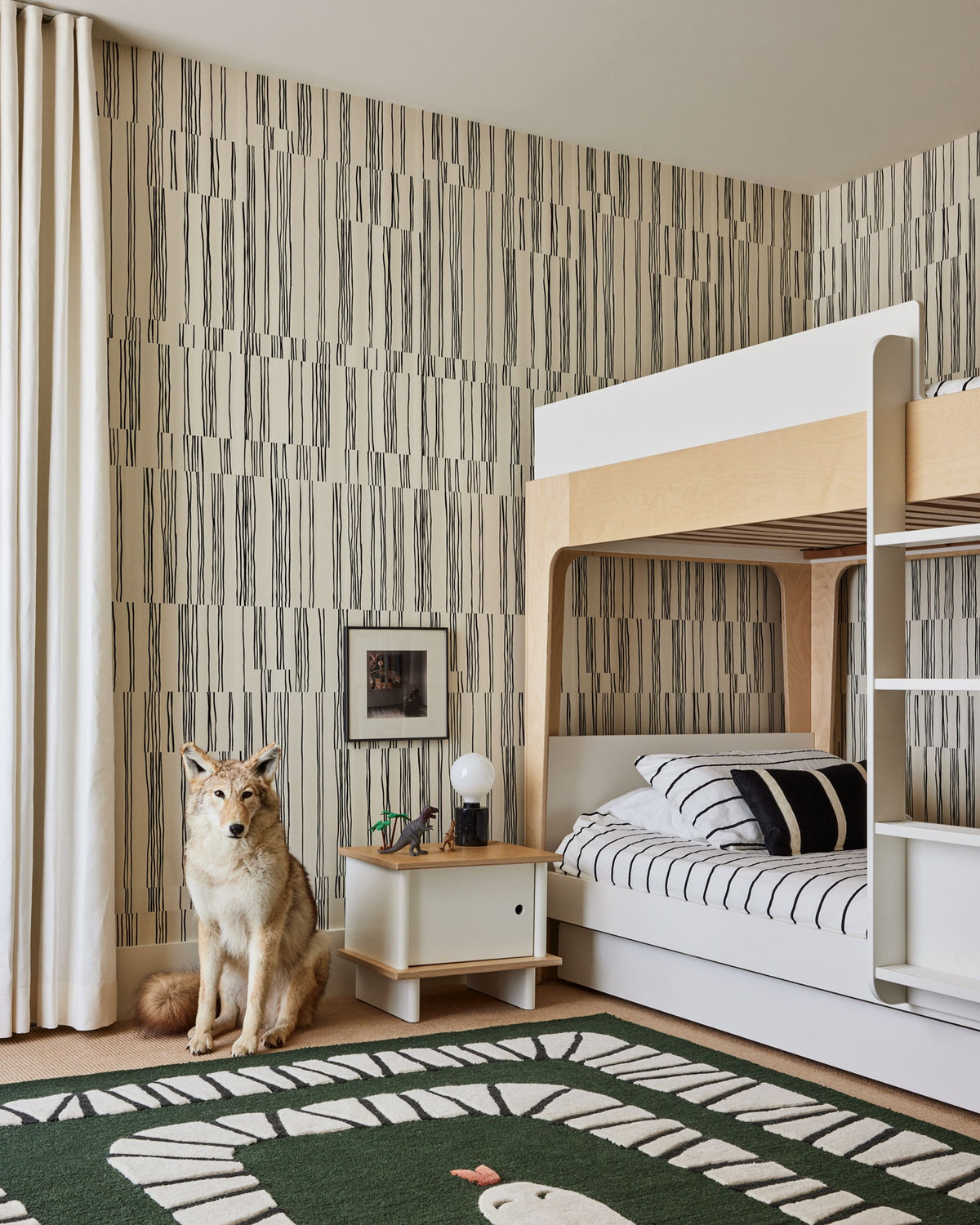 A styled children's bedroom featuring a white and light wood bunk bed, a wolf figurine on the floor, and a green rug with a dinosaur design. The walls are covered in vertical striped wallpaper in beige, gray, and brown tones.