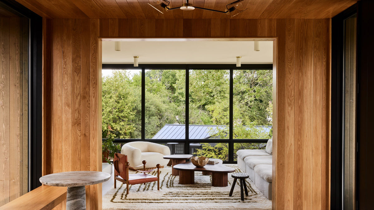 A modern interior view looking out through large black-framed windows onto a lush green outdoor patio and trees, with a light beige sofa, white armchair, wooden coffee tables, and warm wood-paneled walls.