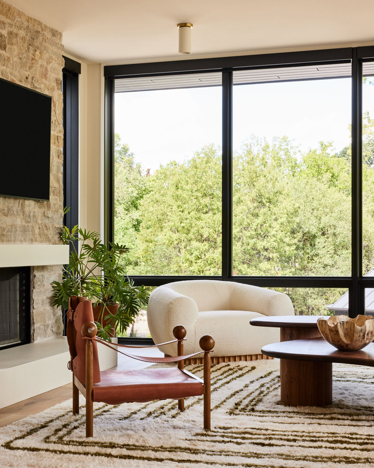 A modern living room interior with a large window overlooking green trees, featuring a cream boucle armchair, a reddish-brown leather accent chair, a patterned rug, and a stone fireplace.