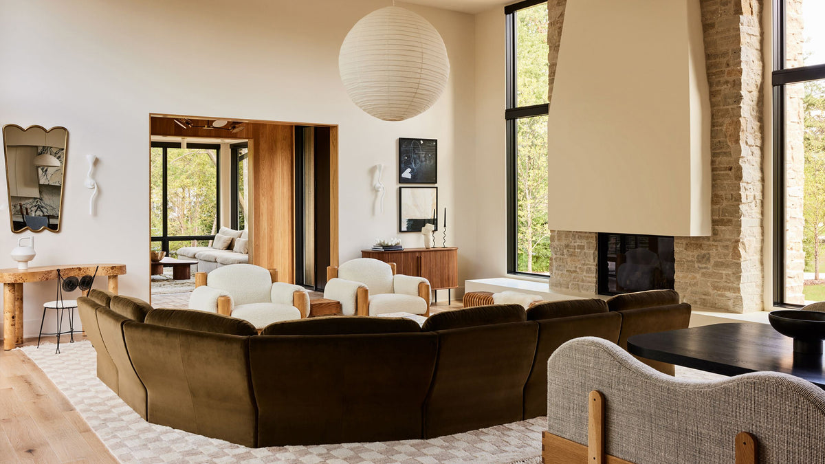 A modern living room featuring a curved sectional sofa upholstered in dark brown velvet, set on a patterned rug in front of a stone fireplace and large windows.