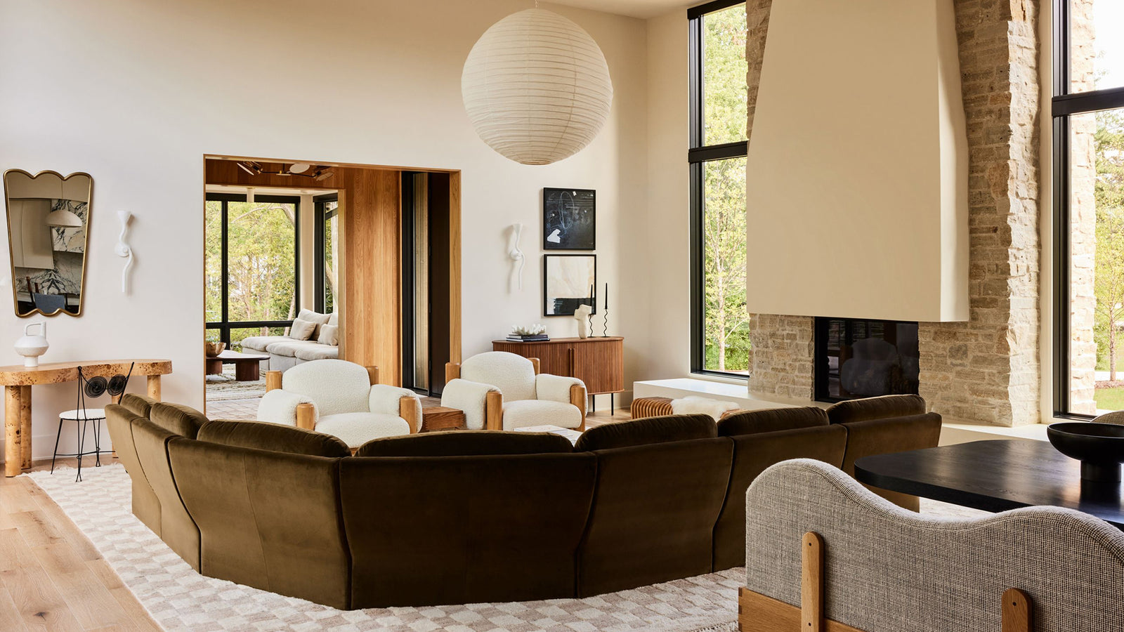 A modern living room featuring a curved sectional sofa upholstered in dark brown velvet, set on a patterned rug in front of a stone fireplace and large windows.