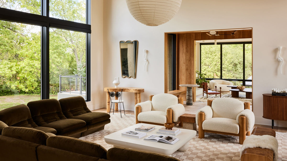 A modern living room interior featuring a dark brown velvet sofa, two cream armchairs with light wood frames, a white coffee table, and large windows looking out to green trees.