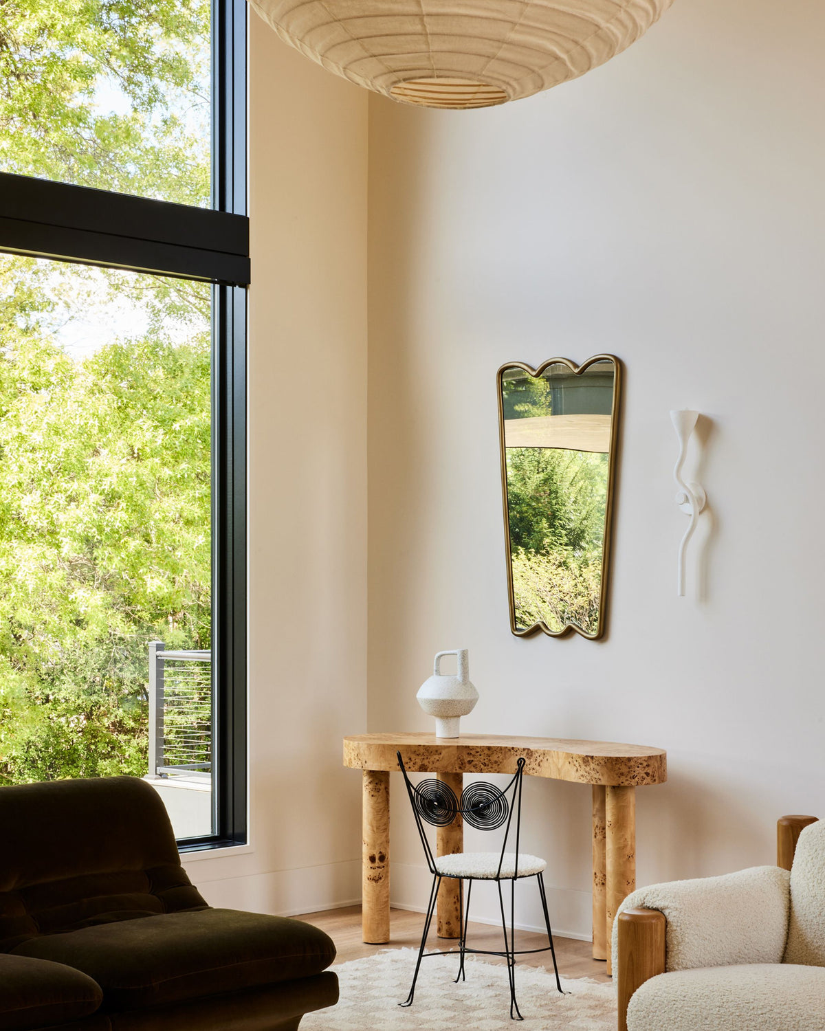 A styled interior corner featuring a tall, scalloped gold-framed wall mirror hanging on a warm beige wall above a rustic wood console table. A white ceramic vase sits on the table, which is paired with a black wire chair with circular back cushions.