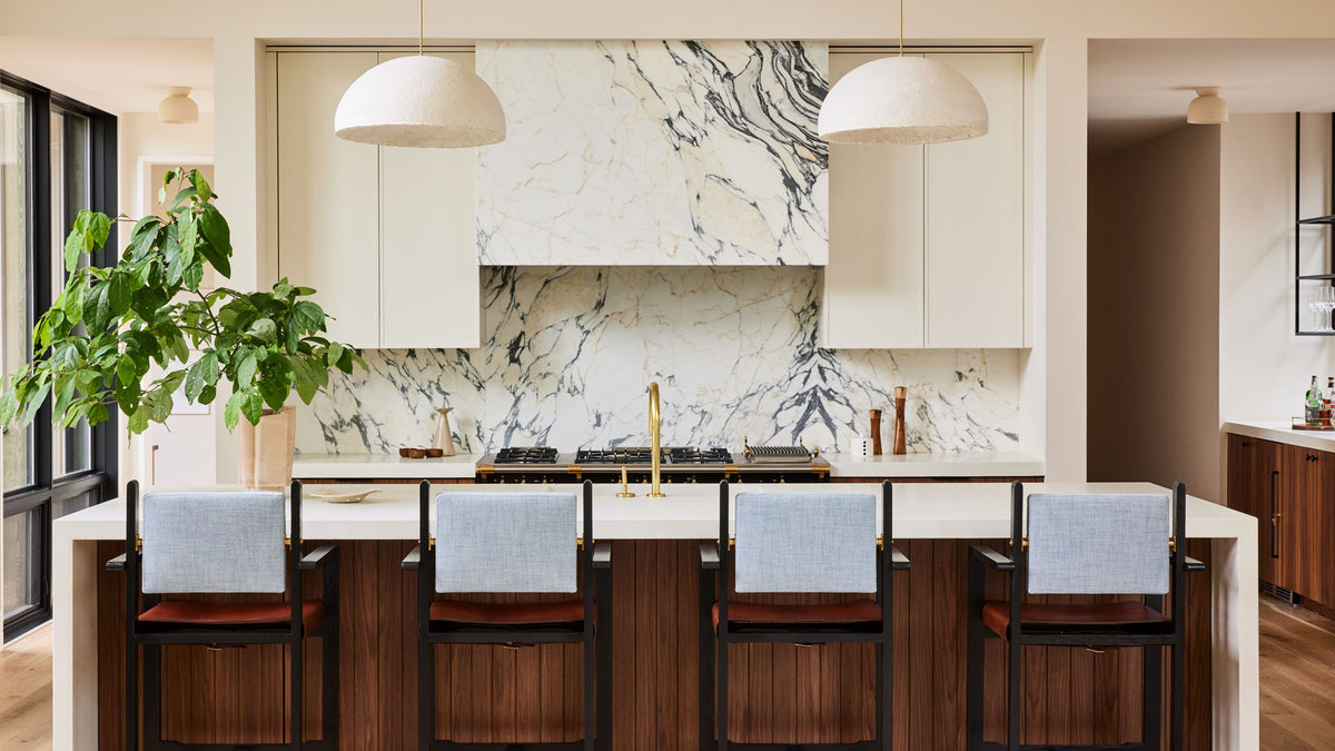 A modern kitchen interior with cream cabinetry, a white marble backsplash, a large white range hood, a central island, and five wooden bar stools with light blue upholstered backs, under two dome pendant lights.
