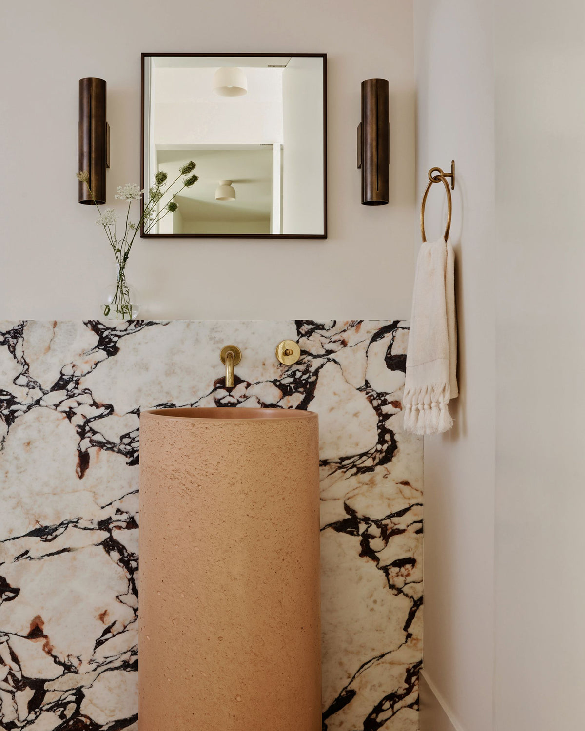 A modern bathroom corner featuring a freestanding terracotta pedestal sink in front of a white marble wall with black veining. Above the sink is a rectangular mirror with a thin black frame, flanked by two dark cylindrical wall sconces.