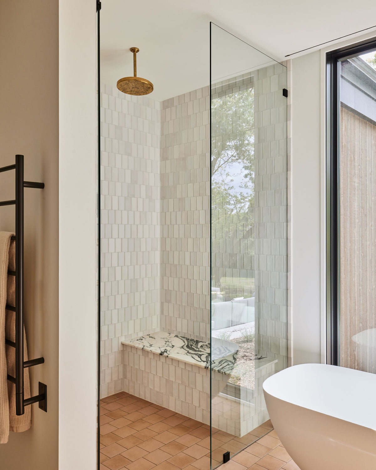 A modern bathroom interior featuring a glass shower enclosure around a tiled walk-in shower with a built-in bench, a freestanding white bathtub edge, and a black towel rack on the left.