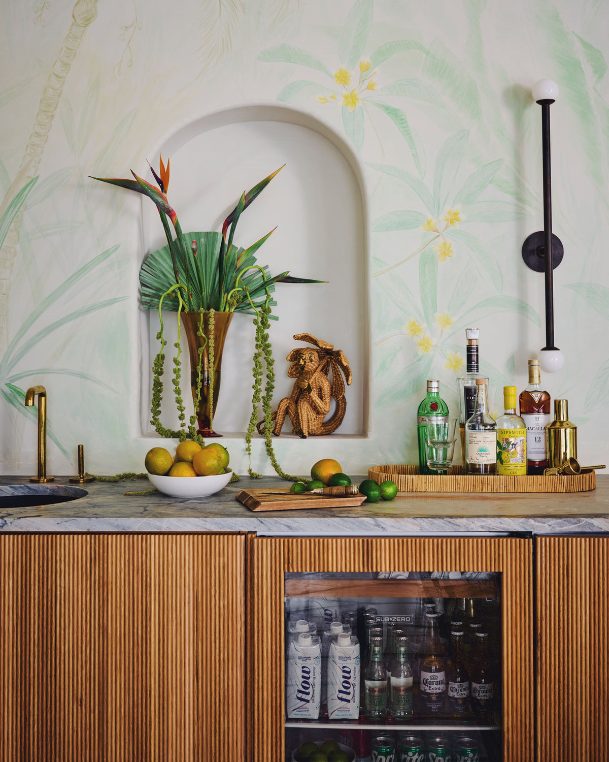 A styled bar counter with a marble top,  wood cabinetry, and a glass beverage refrigerator. A white bowl of lemons and limes sits on the counter beside a wooden tray holding assorted bottles, including a green bottle and a gold-toned bottle.