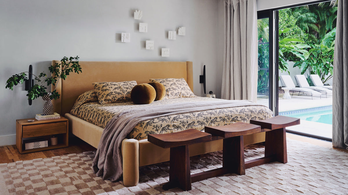 A styled bedroom interior a tan upholstered headboard and frame, matching bench, wooden nightstand, and a large window looking out to a poolside patio and lush greenery.