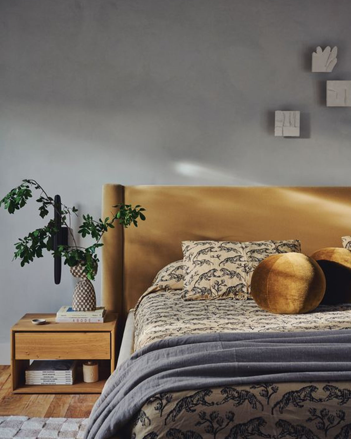 A styled bedroom scene featuring a bed with an upholstered headboard, round gold and black pillows, a wooden bedside table with a plant, and a textured  wall in the background.