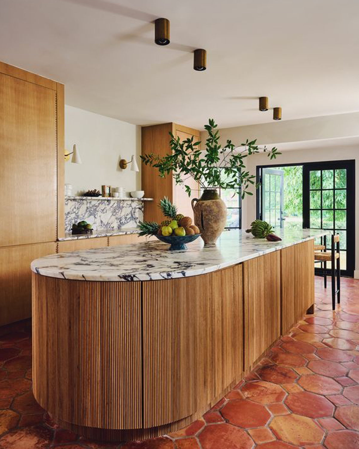 A modern kitchen island with a curved fluted wood cabinet base and a white marble countertop, styled with a large ceramic vase of greenery and fruit. The room features terracotta hexagonal floor tiles, recessed ceiling lights, and a large window in the background.