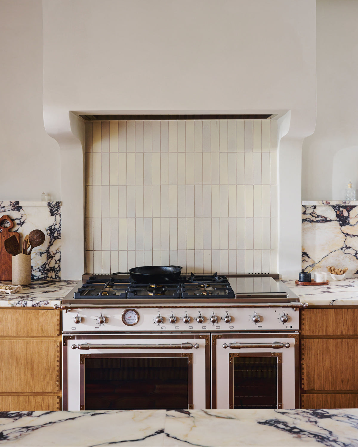 A cream-colored subway tile backsplash installed behind a stainless steel gas range and cooktop, set within a white range hood in a kitchen with marble countertops and wood cabinets.