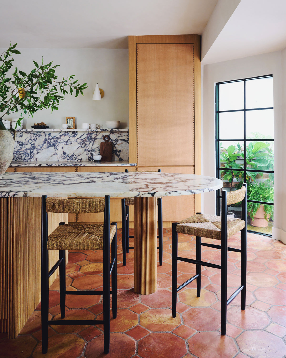 A modern kitchen interior with a marble-topped island, fluted wood base, woven bar stools, terracotta hexagonal floor tiles, and a large window overlooking potted plants.