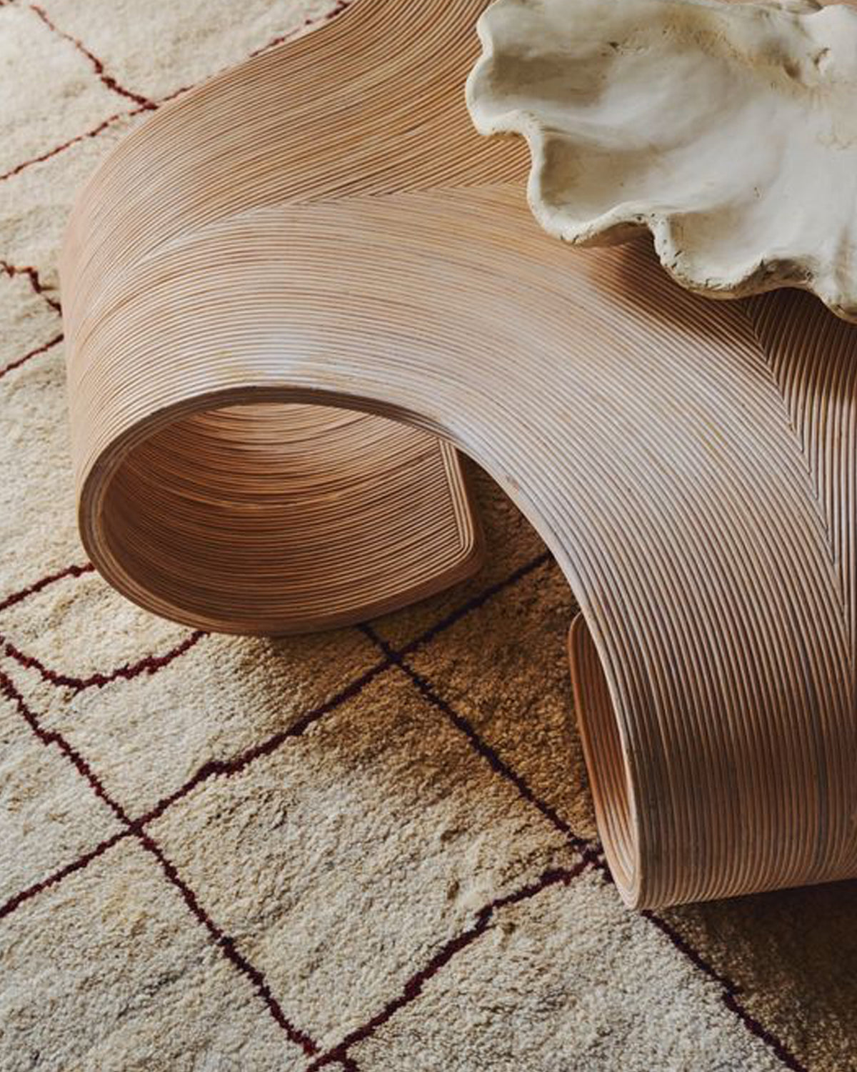 A sculptural, curved wooden side table styled on a beige patterned rug next to a white ceramic shell.