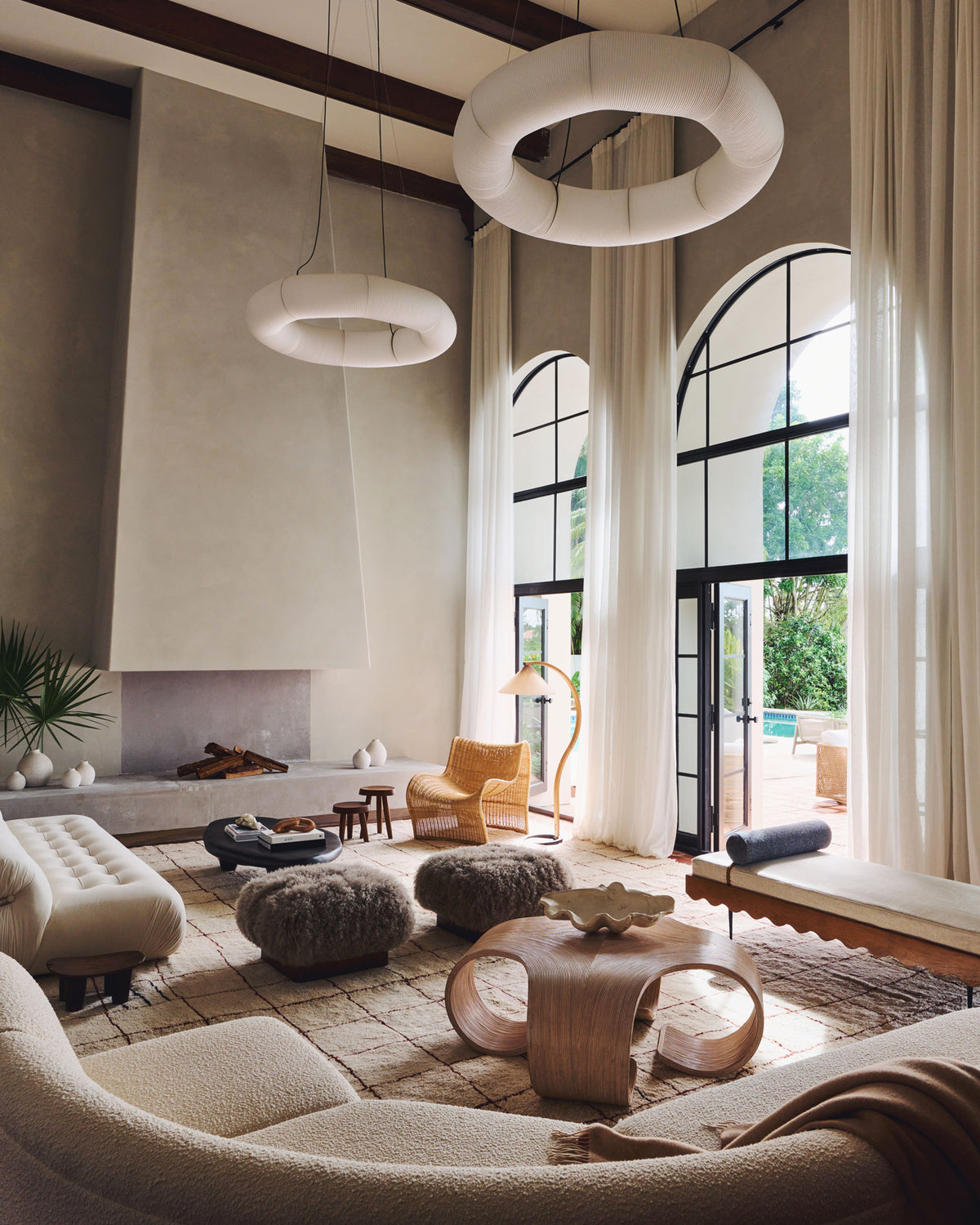 A luxury modern living room featuring a sculptural cream bouclé sofa in the foreground, textured poufs, a curved wooden coffee table, a stone fireplace, and large arched windows with white curtains.