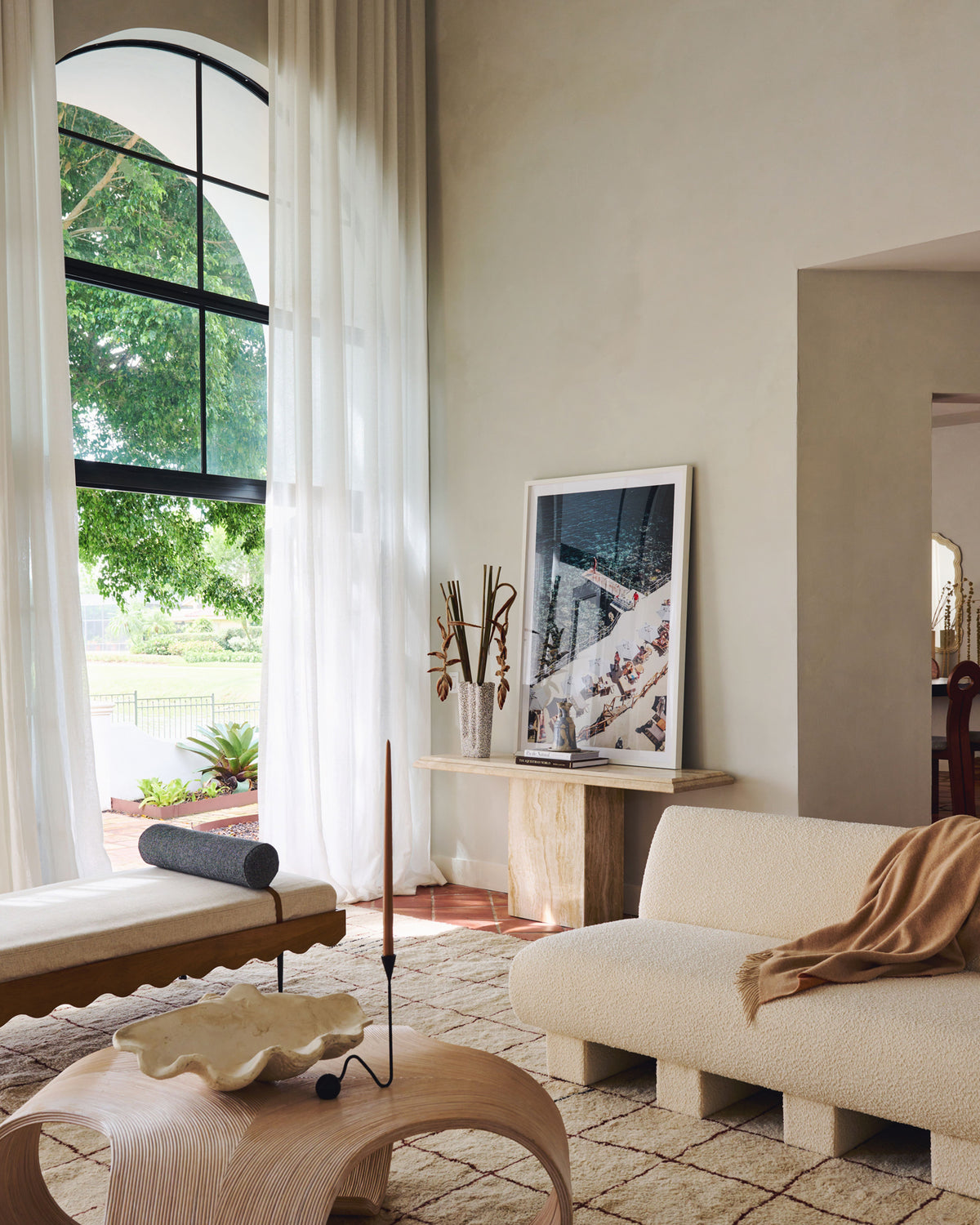 A styled interior room scene with a cream upholstered sofa, a sculptural light wood coffee table, a bench with a dark cushion, a tall window with white curtains, and framed wall art against a warm beige wall.