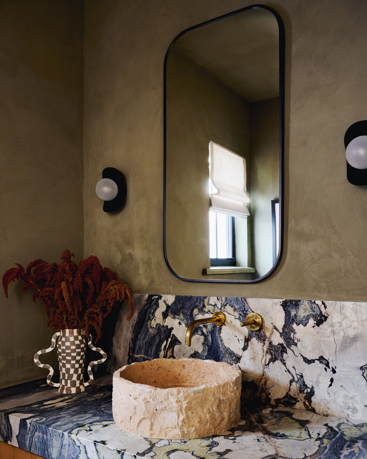 A stone basin sink set into a blue and white veined marble countertop, with a large black-framed wall mirror above and a gold faucet. 
