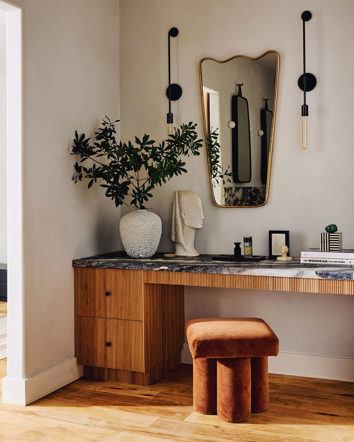 A styled interior corner featuring a wooden vanity desk with a dark marble top, gold-framed mirror, black wall sconces, a white textured vase with green branches, a white sculptural bust, and a terracotta velvet stool on a light wood floor.