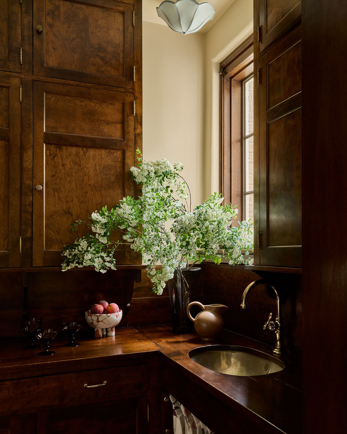 A kitchen butler pantry corner with dark wood cabinets, a wooden countertop, a brass sink, and a large vase filled with white flowers on the counter.