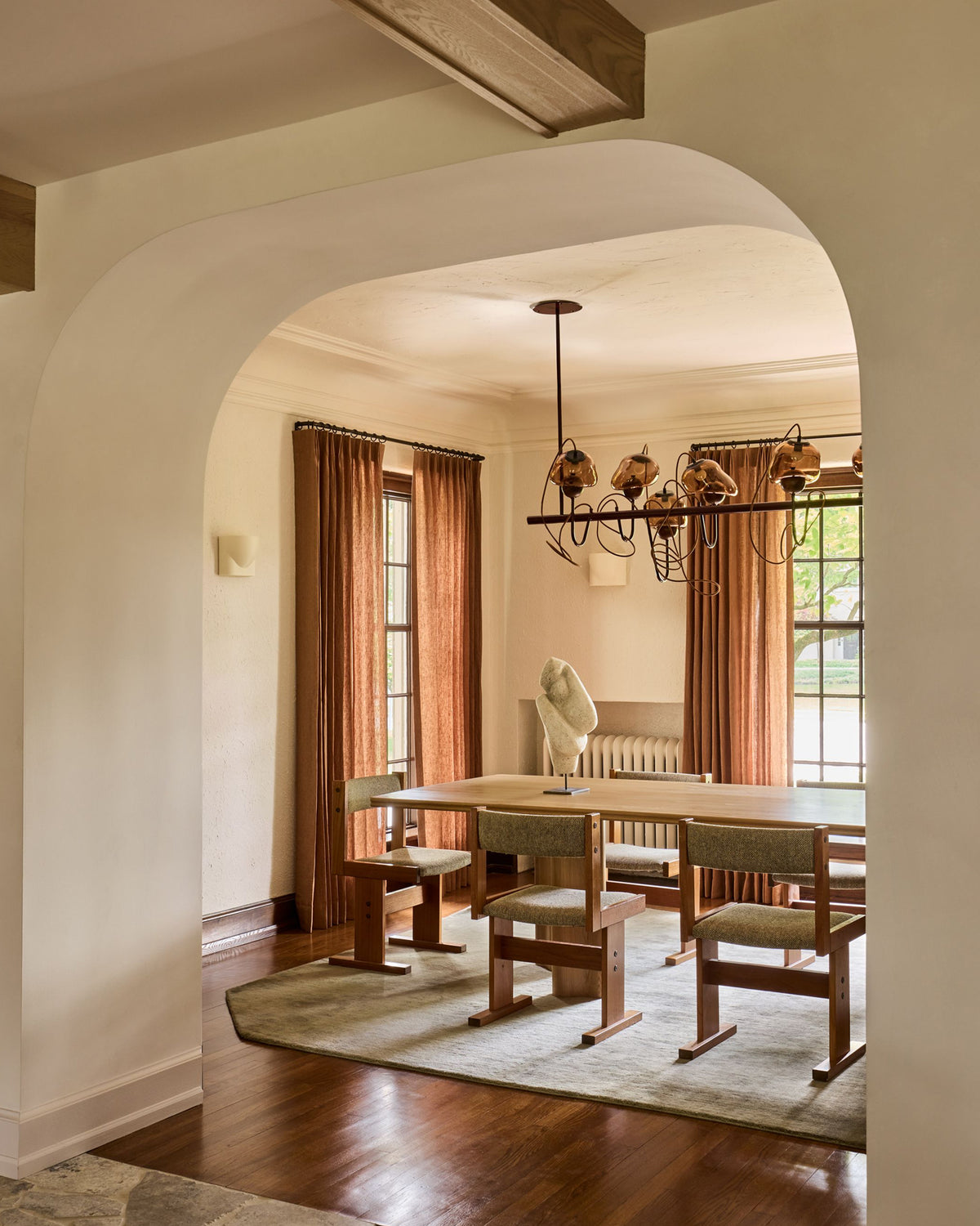 A dining room viewed through a large white archway, featuring a light wood dining table, four wooden chairs with light upholstery, a beige rug, rust-colored curtains, and a black chandelier.