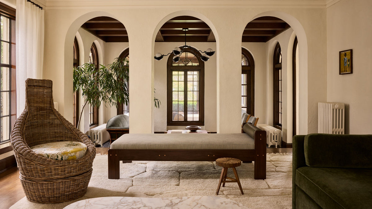 A styled interior room with arched openings, featuring a woven wicker accent chair with a cushion, a low wooden daybed with a light upholstered seat, a small wooden stool, a green sofa edge, and plants in a Mediterranean-inspired room setting.