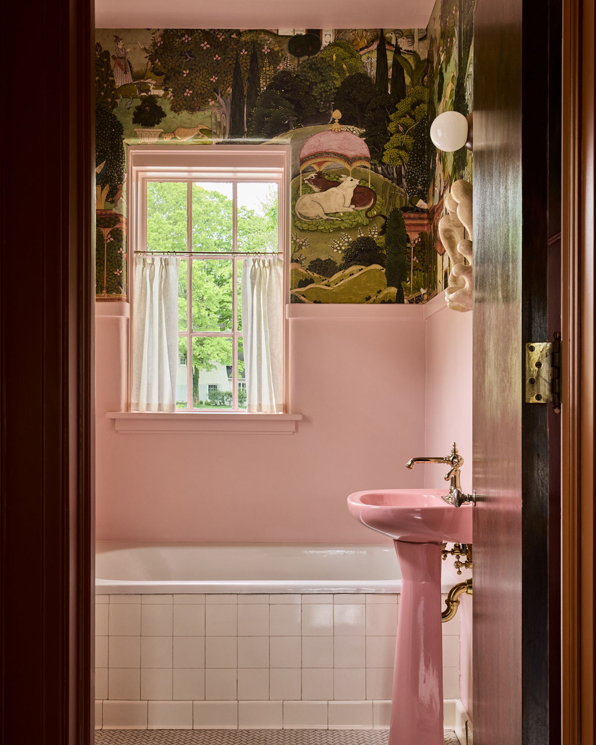 A pink bathroom interior viewed through an open dark wood doorway, featuring a pink pedestal sink, white tiled tub, window with white curtains, and a whimsical forest-patterned wallpaper.