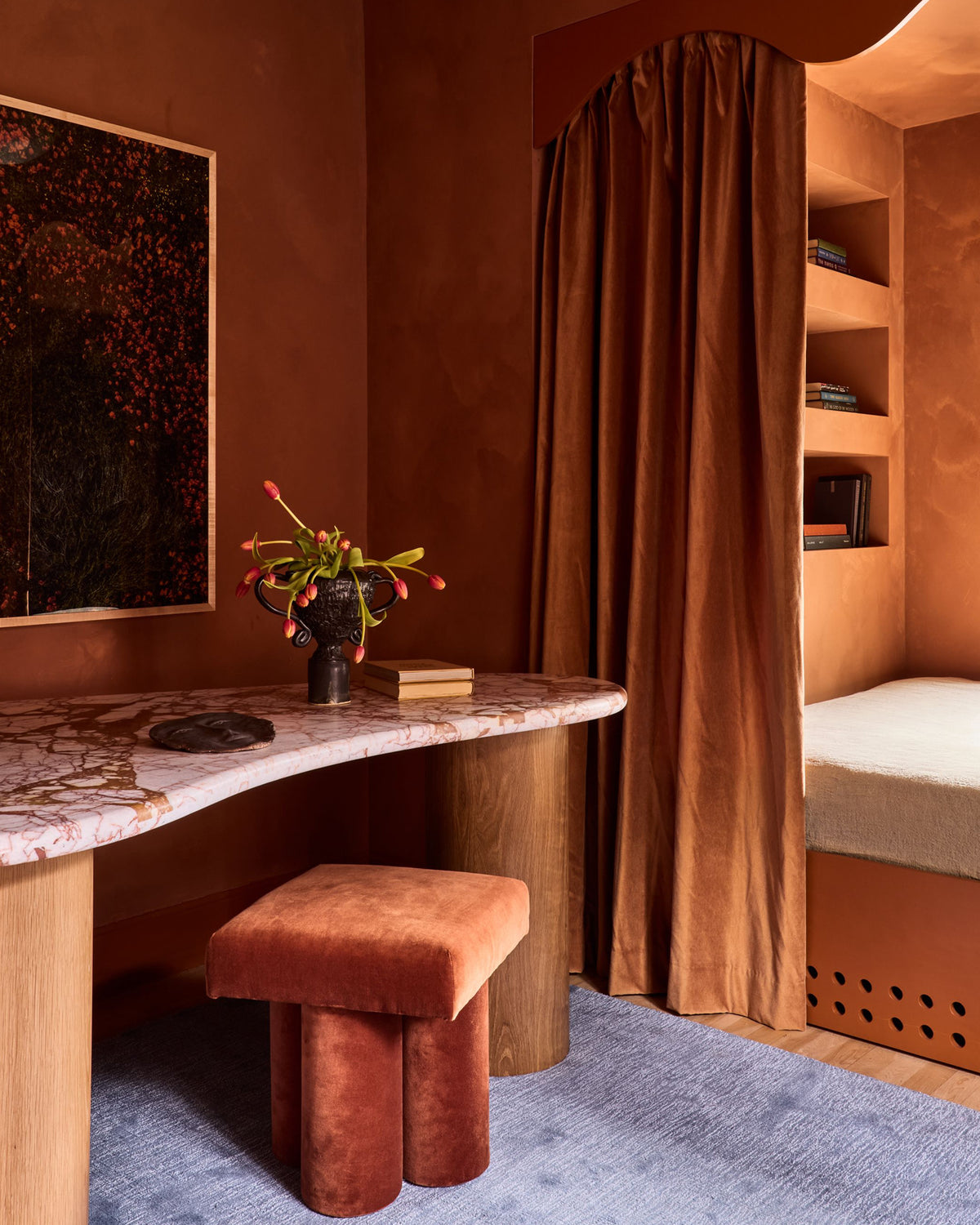A warm, earthy-toned bedroom interior featuring a curved desk with a marble-patterned top, a matching velvet stool, a brown velvet curtain, and built-in shelving with a bed visible in the background.