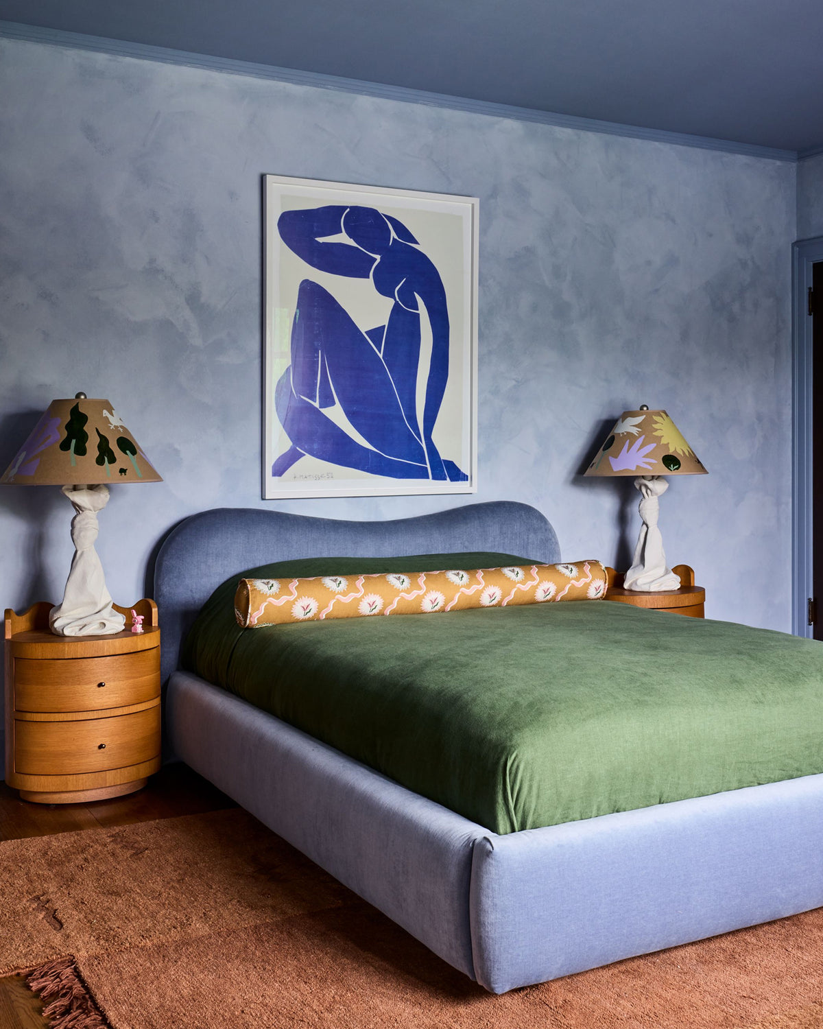A styled bedroom featuring a light blue upholstered bed with a green velvet cover, flanked by two wooden nightstands and sculptural table lamps. Above the bed, a white-framed art print.