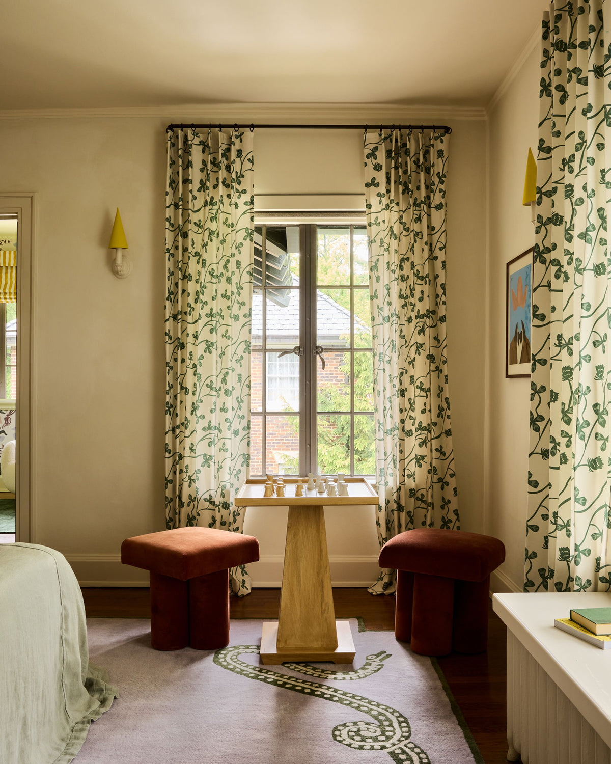 A styled bedroom interior featuring a window dressed with patterned curtains in green vines on a cream background, with two rust-red upholstered ottomans and a small wooden table in front. A bed edge, wall sconces, and a rug are also visible.