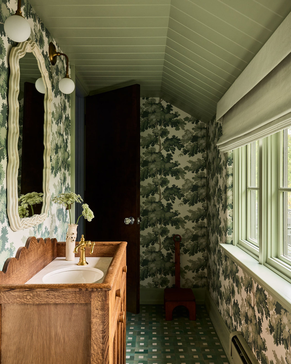 A narrow vintage-style bathroom with green and white floral wallpaper, a wooden vanity with a white sink, a large ornate mirror, a dark wooden door, and a window with a white blind.